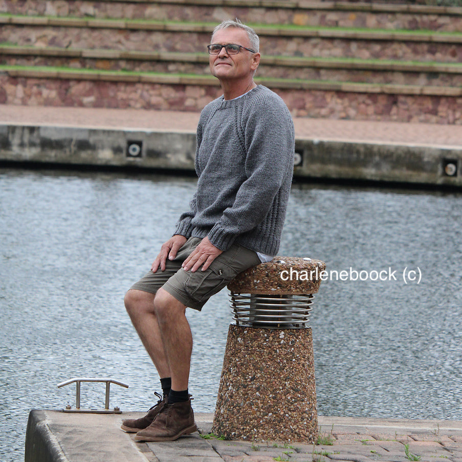 A man sitting on a stone and metal post by the water, wearing a hand-knit gray sweater with raglan sleeves, khaki shorts, dark socks, and brown shoes.