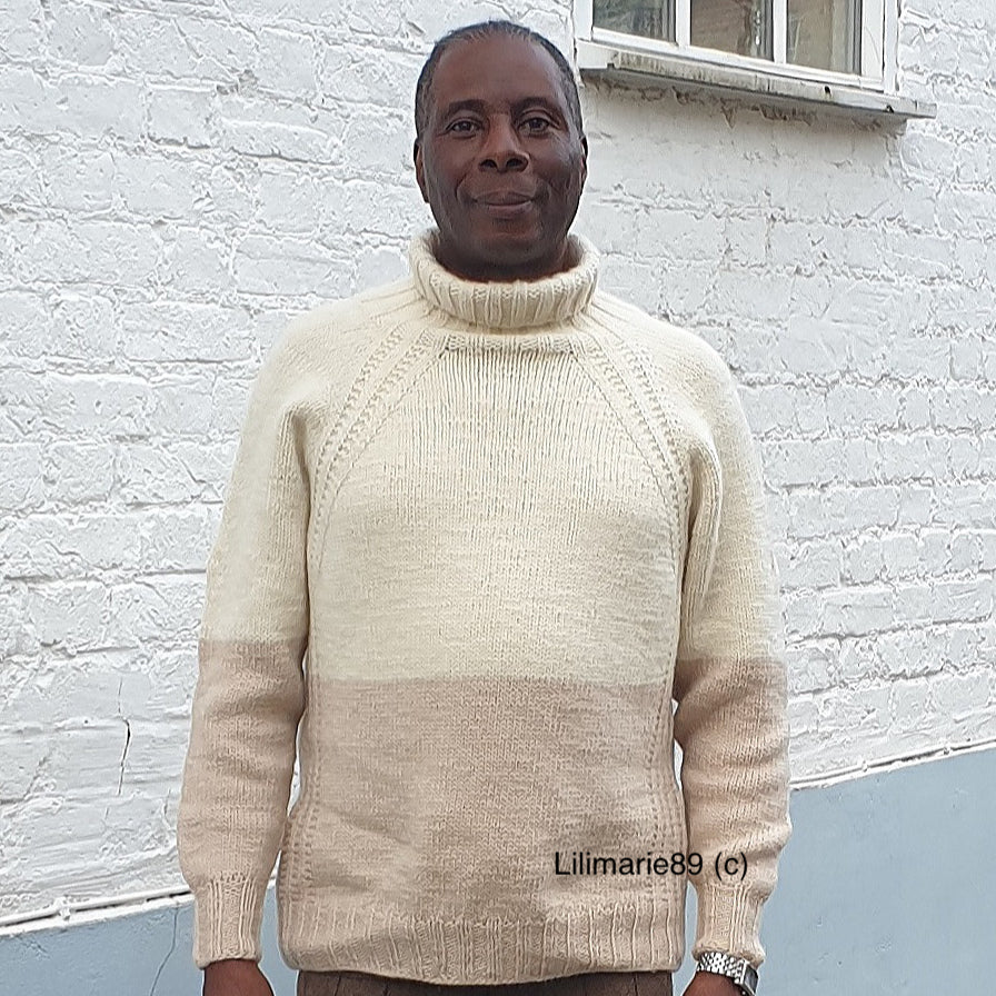 A man standing outdoors against a white brick wall, wearing a hand-knit turtleneck sweater with raglan sleeves, designed in a two-tone color block style of cream and beige.