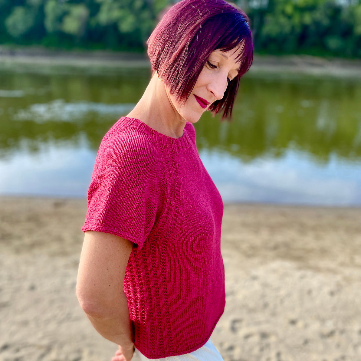 Woman with short red hair wearing a bright pink short-sleeved knit sweater standing by a river.
