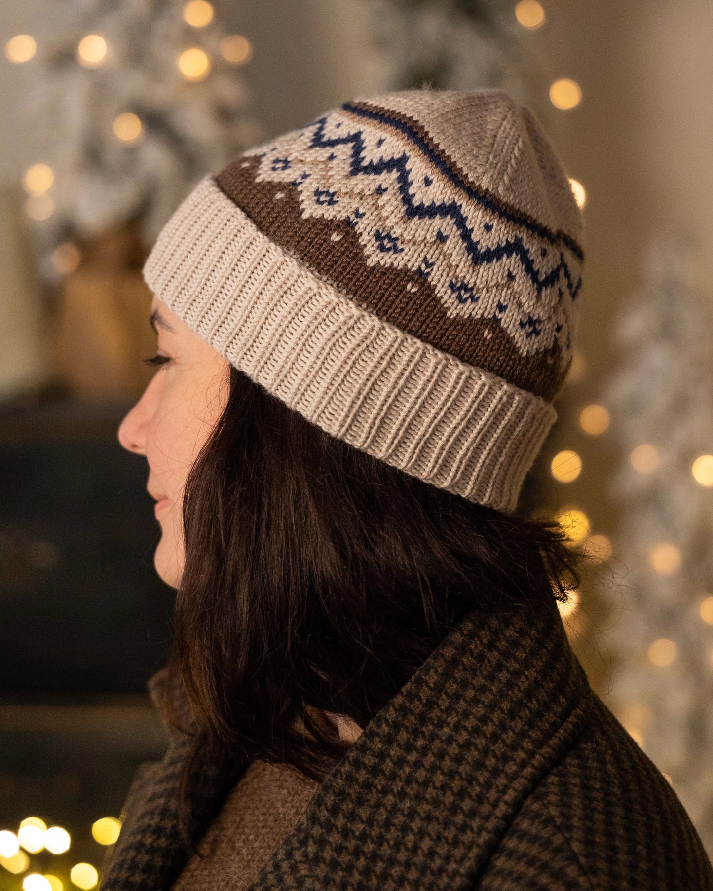 Person wearing a knitted beanie with a pattern in a warm, blurred indoor setting.