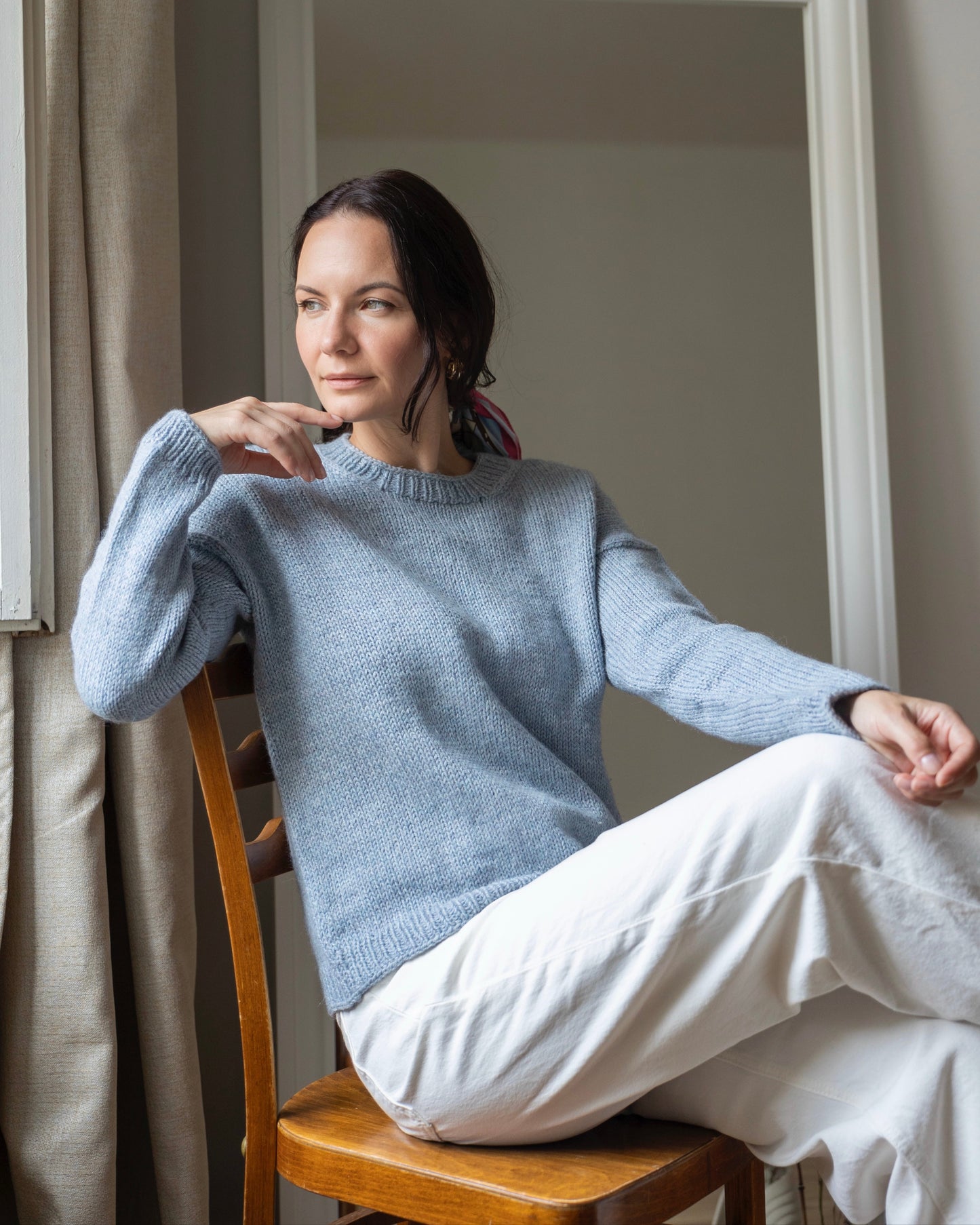 Woman sitting on a wooden chair near a window, wearing a light blue hand-knitted sweater and white pants.