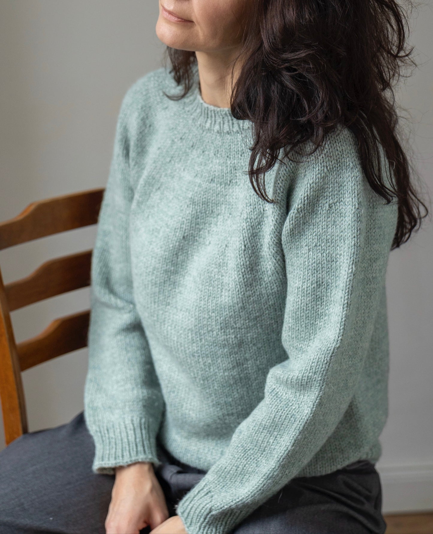 Woman wearing a light green sweater sitting on a wooden chair against a plain wall.