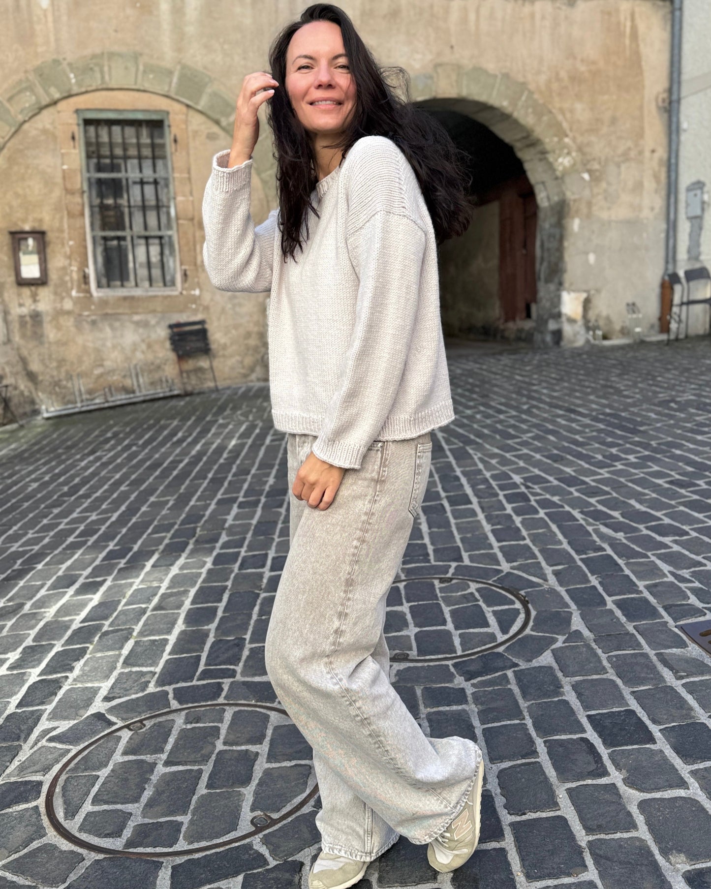 Julia Piro wearing a light beige hand-knit sweater paired with wide-leg jeans and sneakers, standing on a cobblestone street in front of a stone archway.