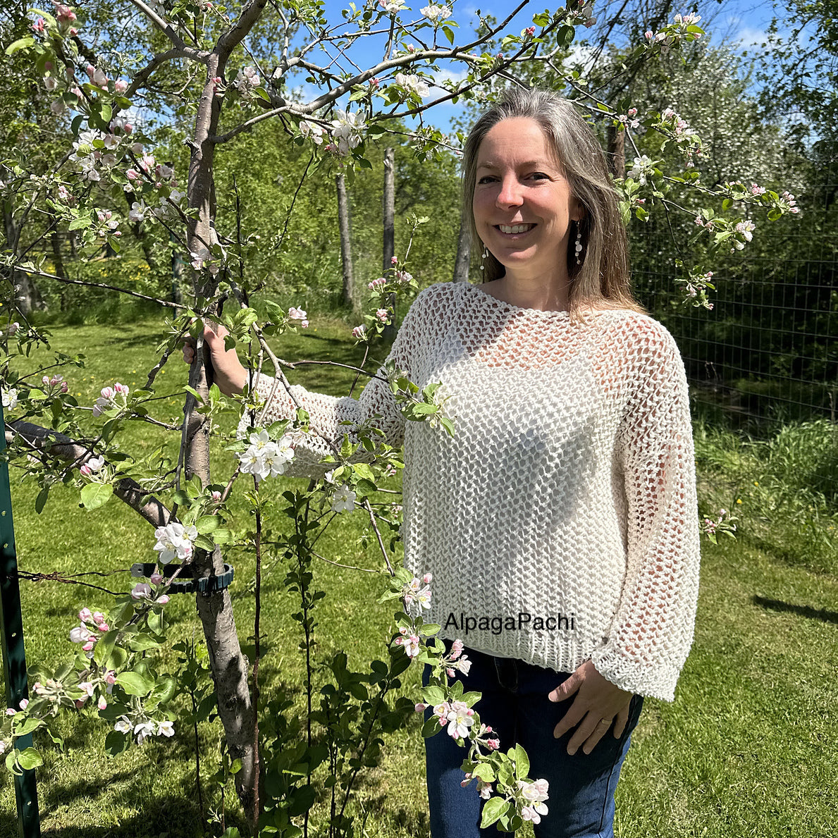 Woman standing outdoors among blossoming trees, wearing the Halcyon Lace Sweater in cream.