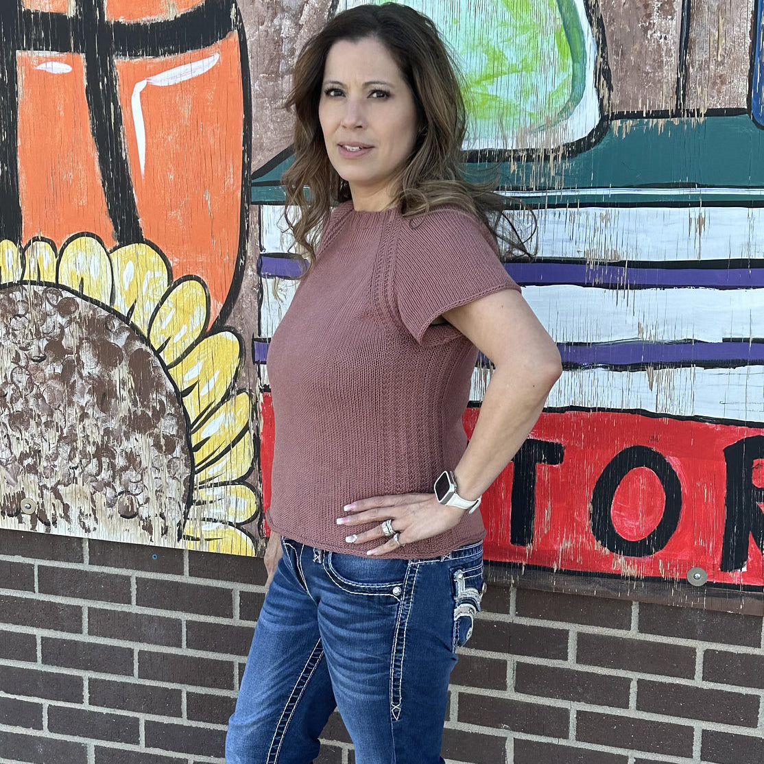 Woman wearing a short-sleeved dusty rose knit sweater paired with jeans standing in front of a colorful mural.