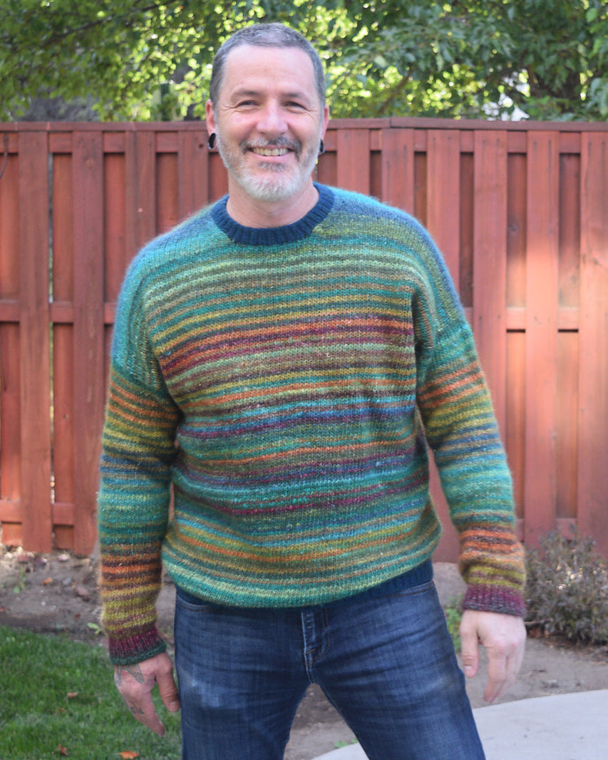 Man smiling and wearing a colorful striped knit sweater with shades of green, blue, orange, and burgundy, standing outdoors by a wooden fence