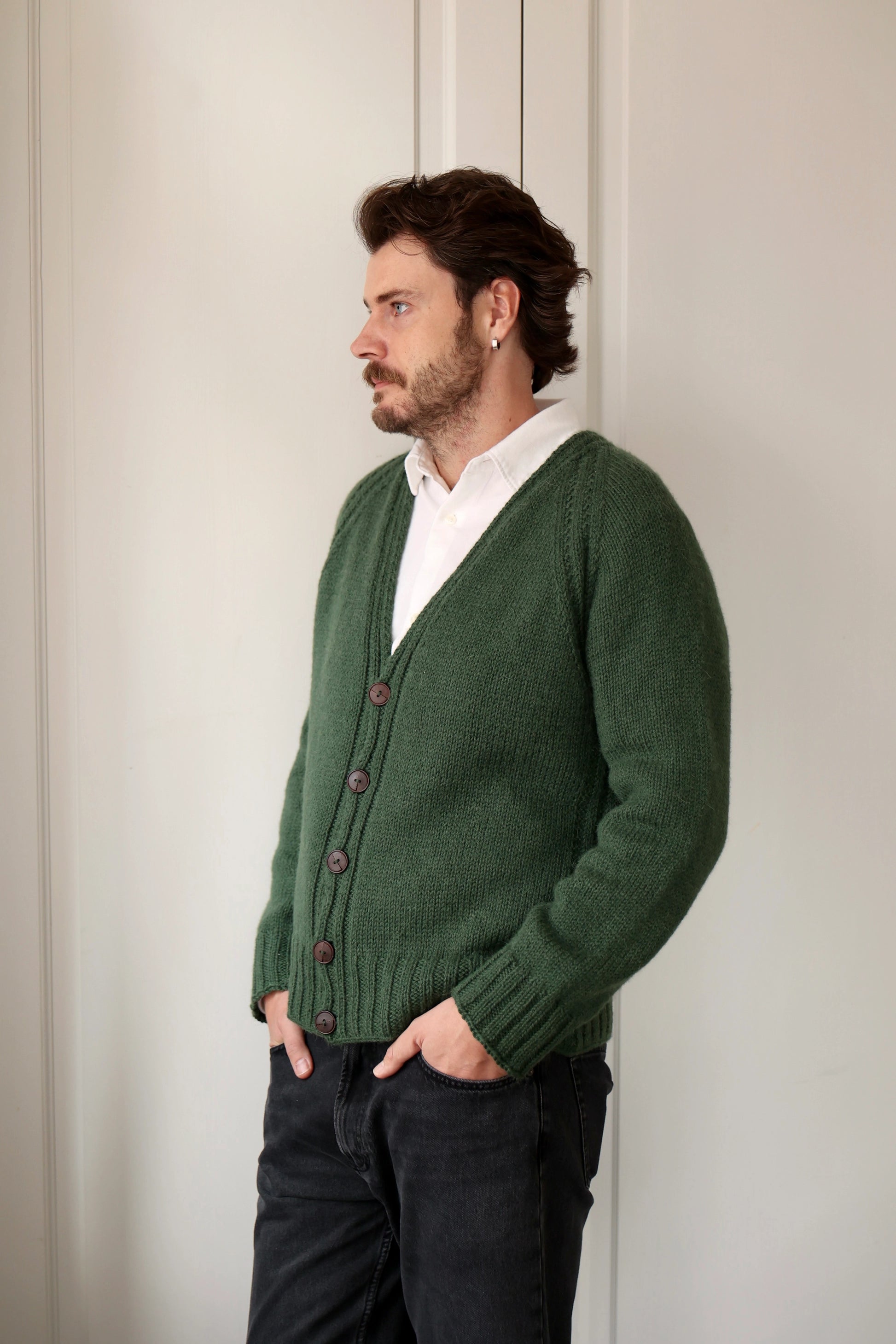 Man wearing the Mellow Mood Cardigan in forest green, standing with hands in pockets and looking to the side, styled with a white shirt and dark jeans.