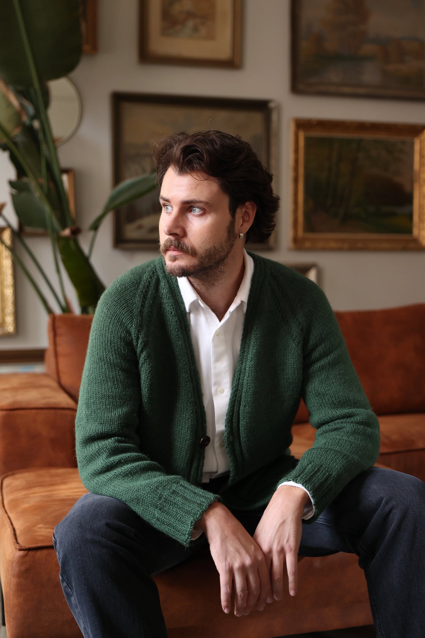 Man wearing the Mellow Mood Cardigan in forest green, seated on a brown sofa in a cozy room with framed artwork and plants in the background.