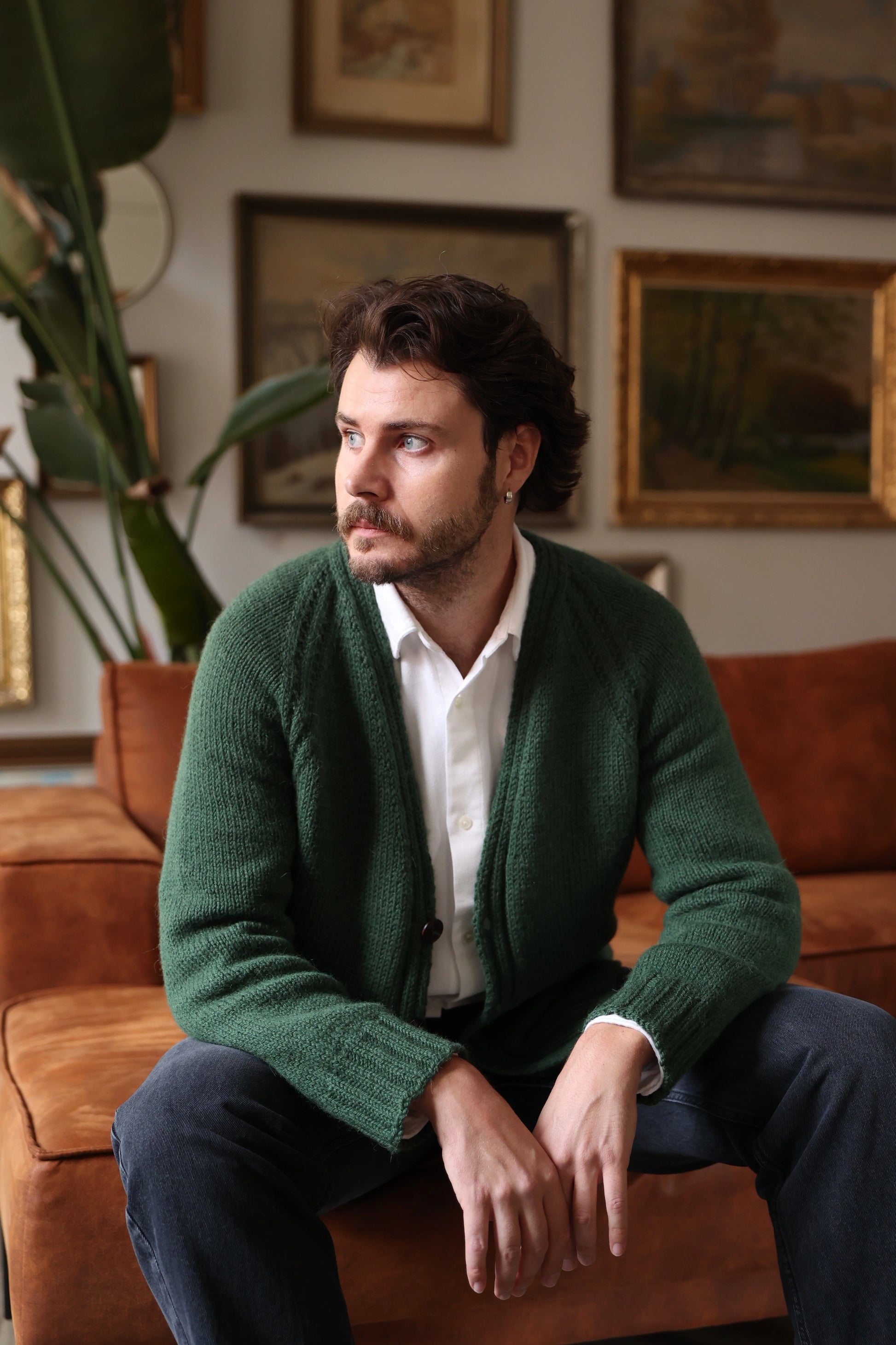Man wearing the Mellow Mood Cardigan in forest green, seated on a brown sofa in a cozy room with framed artwork and plants in the background.