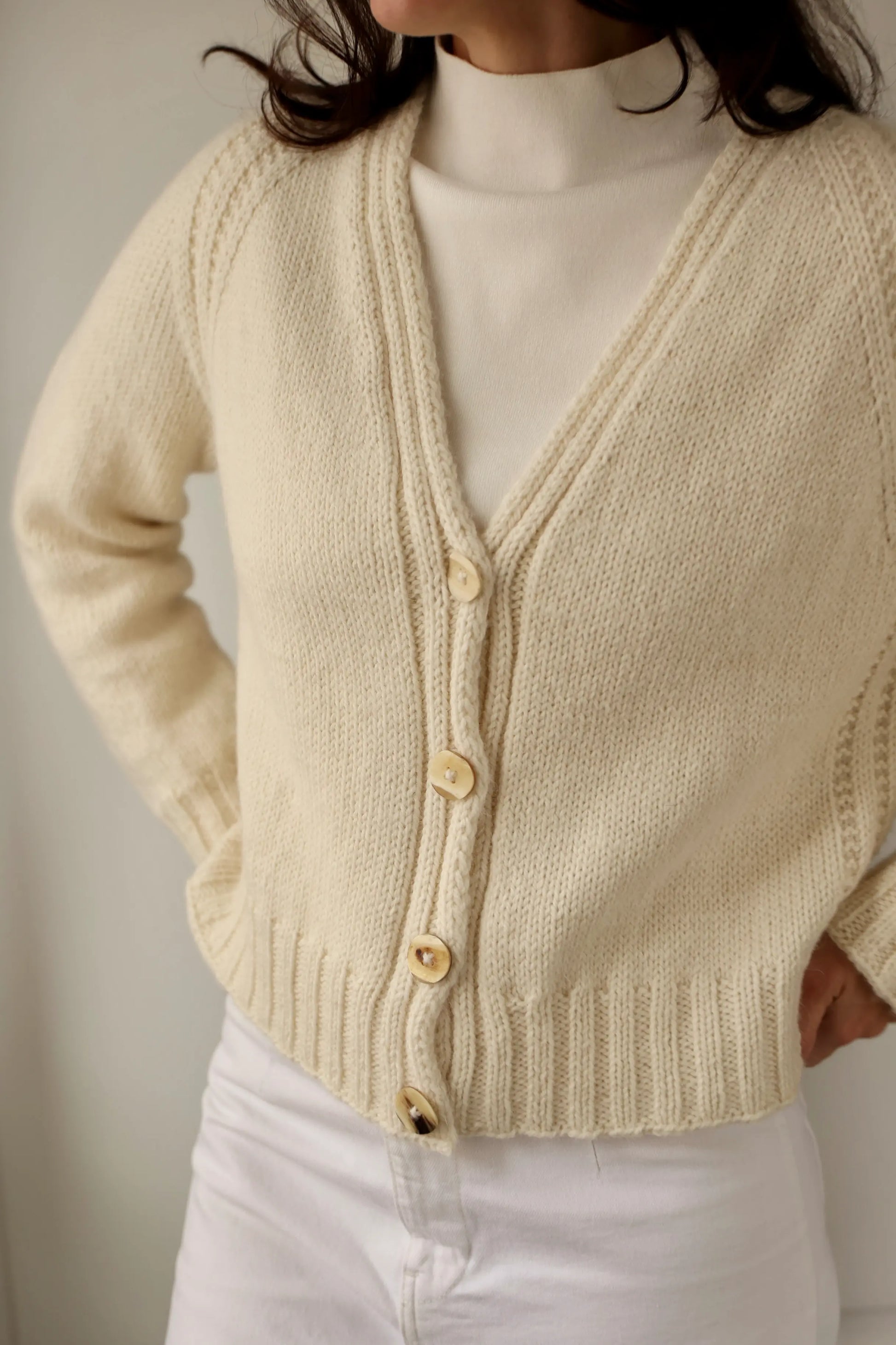 Close-up of a woman wearing the Mellow Mood Cardigan in cream, styled over a white turtleneck and paired with white pants.