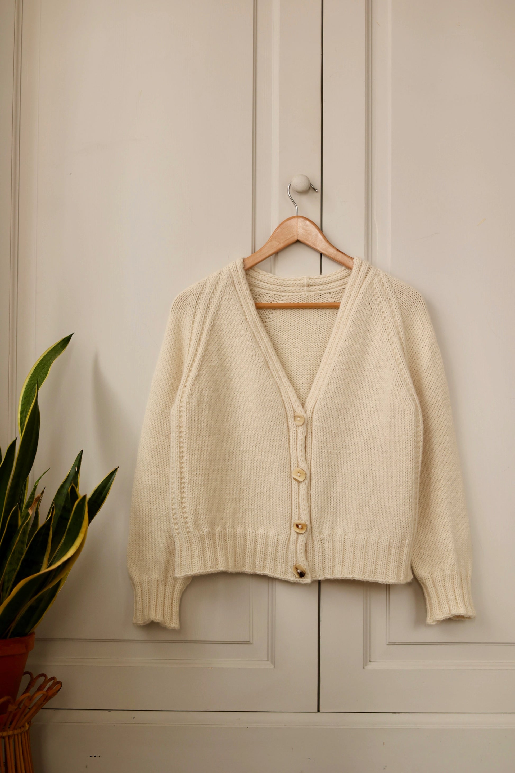 The Mellow Mood Cardigan in cream displayed on a wooden hanger against a light-colored cabinet, with a potted plant beside it.