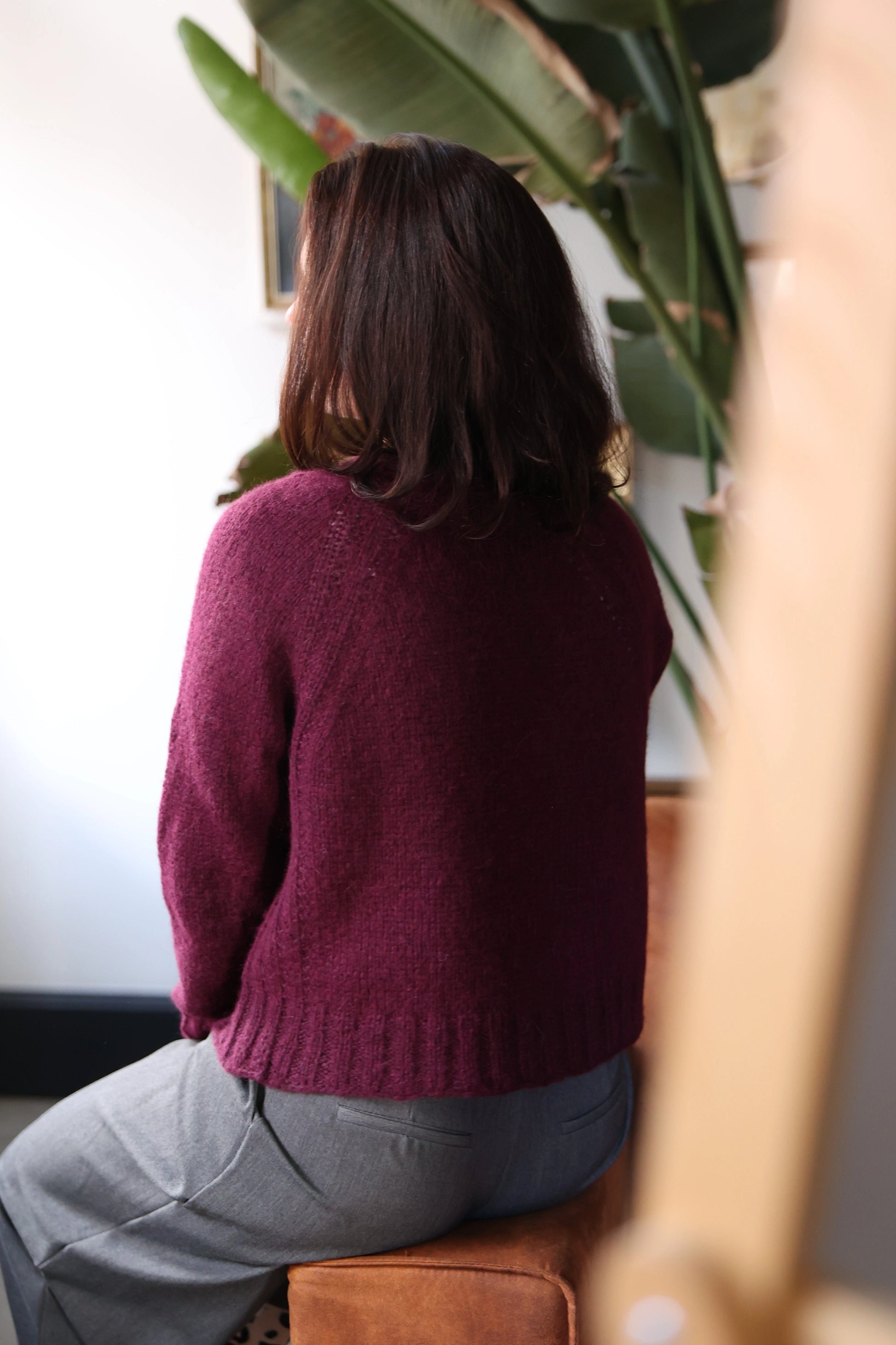 Back view of a woman wearing the Mellow Mood Cardigan in wine color, sitting on a brown sofa in a softly lit room with green plants in the background.