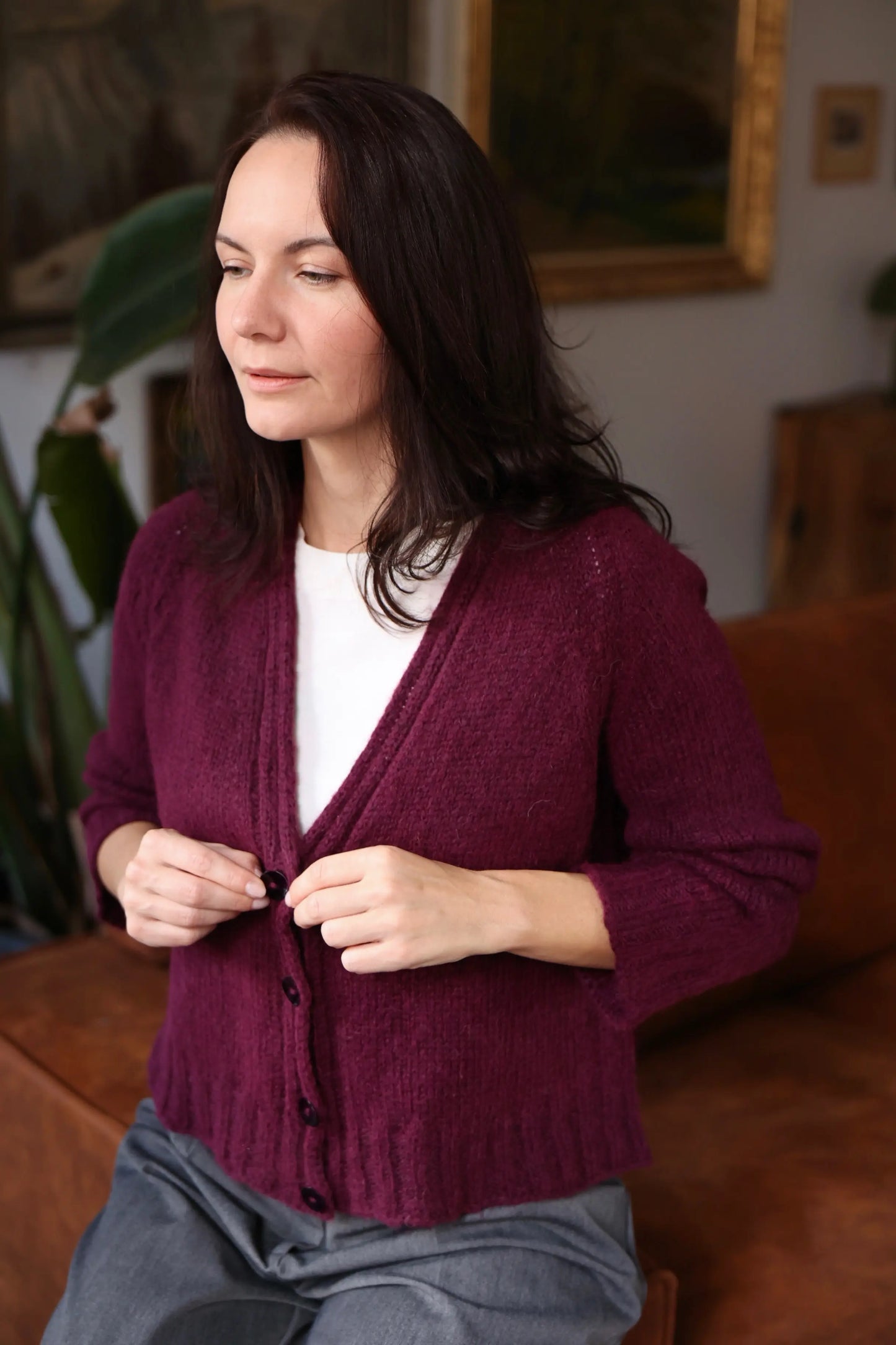 Woman wearing the Mellow Mood Cardigan in a deep wine color, seated on a brown sofa in a softly lit room, buttoning the cardigan over a white top.