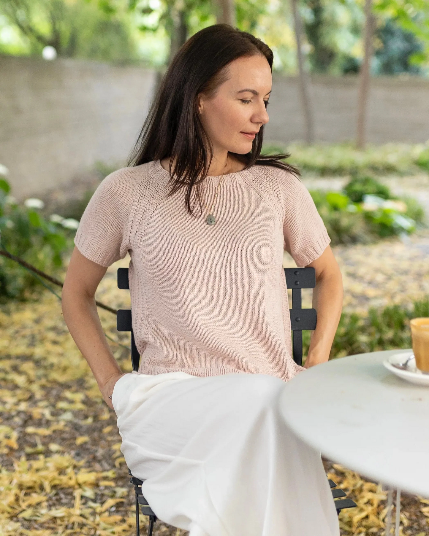 Woman sitting outdoors in a garden setting wearing a light pink sweater and white pants.