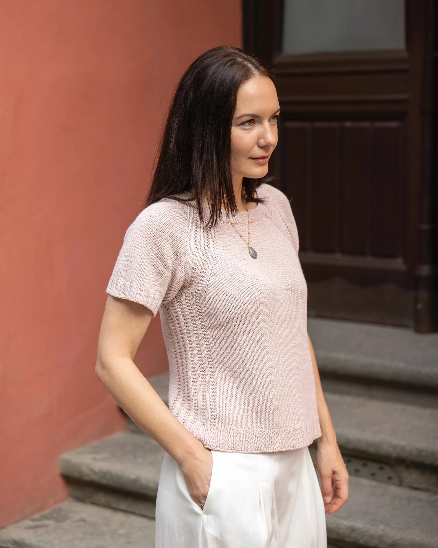 Woman wearing a light pink knitted sweater and white pants standing on steps.
