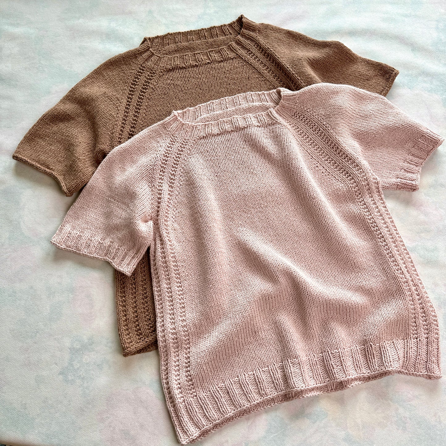 Two knitted Mellow Mood Tops laid flat one in soft brown and one in light pink both featuring short sleeves ribbed edges and textured raglan details.