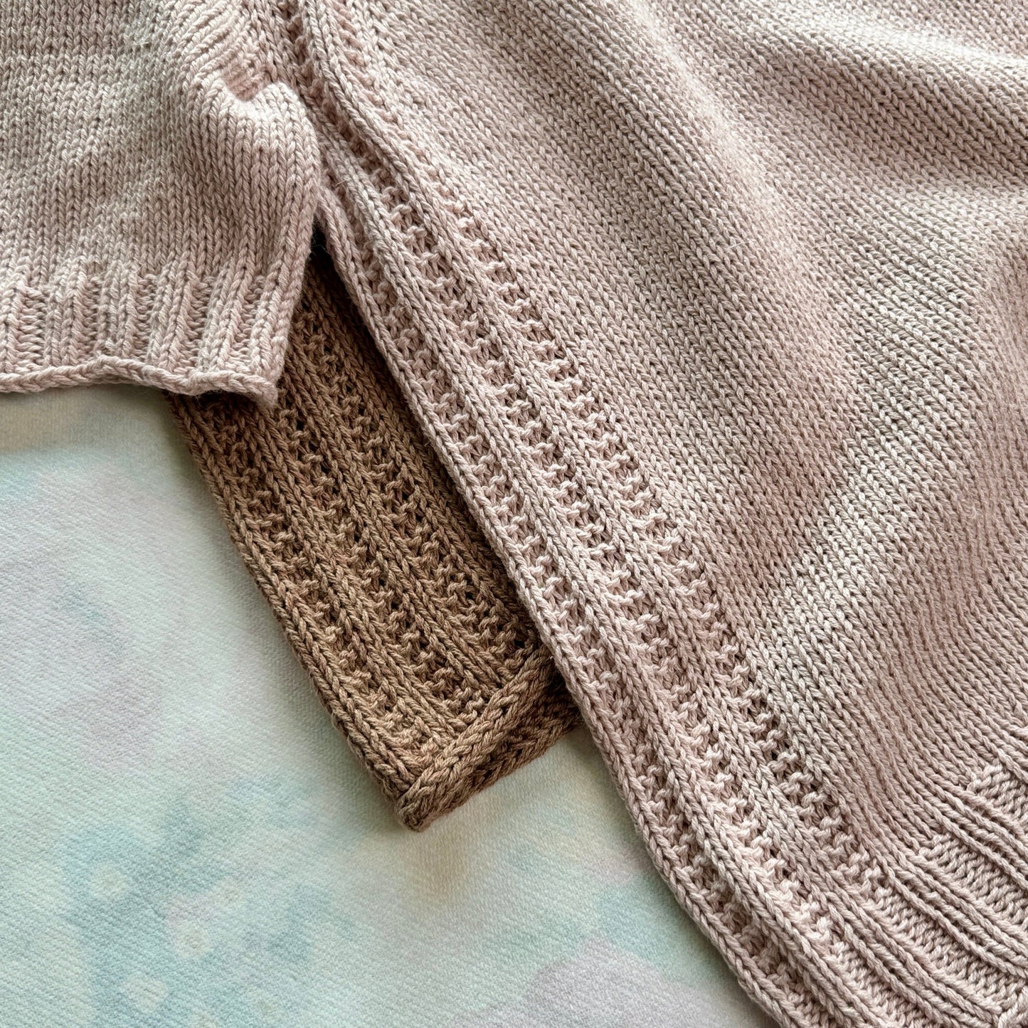 Close up of two knitted Mellow Mood Tops showing the sleeve and side details one in soft pink and one in light brown highlighting the textured raglan stitches.