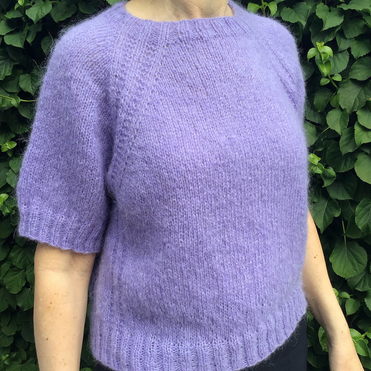 Person wearing a short-sleeved lilac fuzzy knit sweater with ribbed edges standing in front of green foliage.