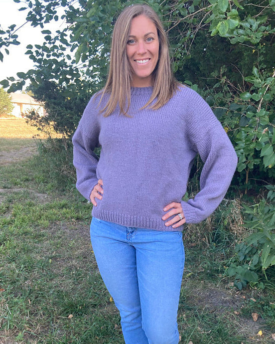 Woman standing outdoors wearing a hand-knit lavender sweater paired with light blue jeans, smiling with hands on hips