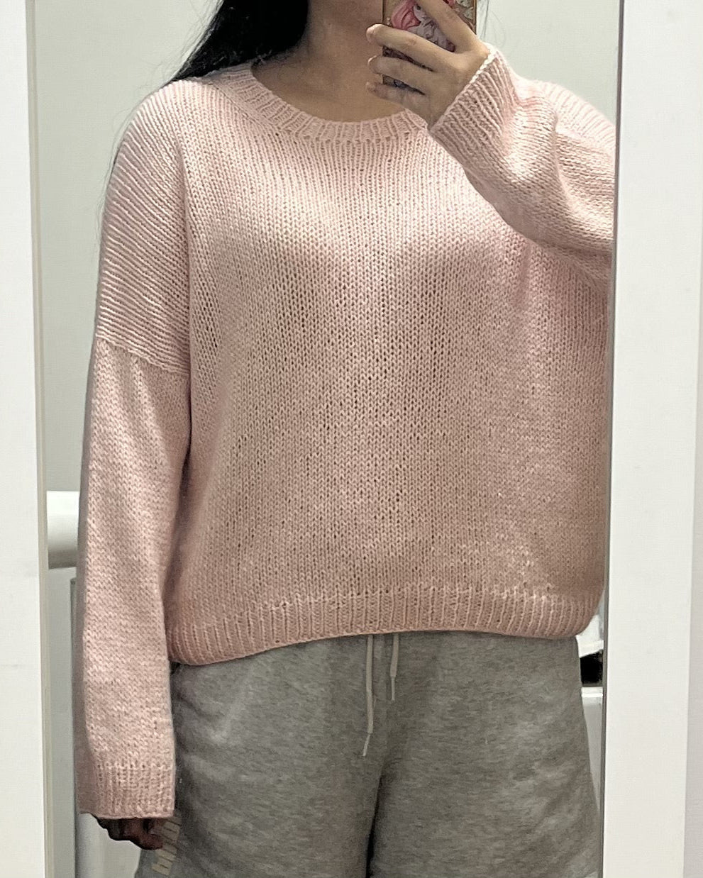 Woman taking a mirror selfie wearing a loose-fitting hand-knit pink sweater paired with gray sweatpants