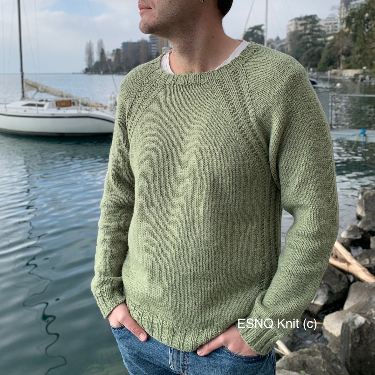 A man stands by a marina with sailboats in the background, wearing sunglasses, jeans, and the His Mellow Mood Sweater in light green.