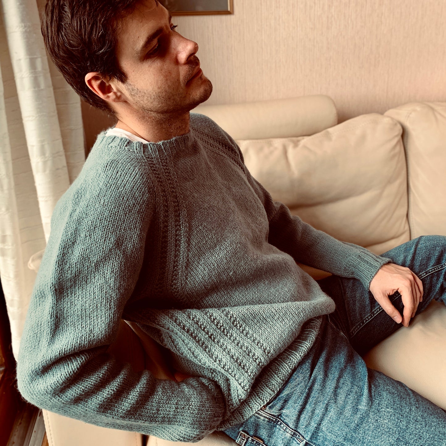 A man relaxing on a beige sofa while wearing a hand-knit gray raglan sweater with textured stitch details along the seams and hem, paired with blue jeans.