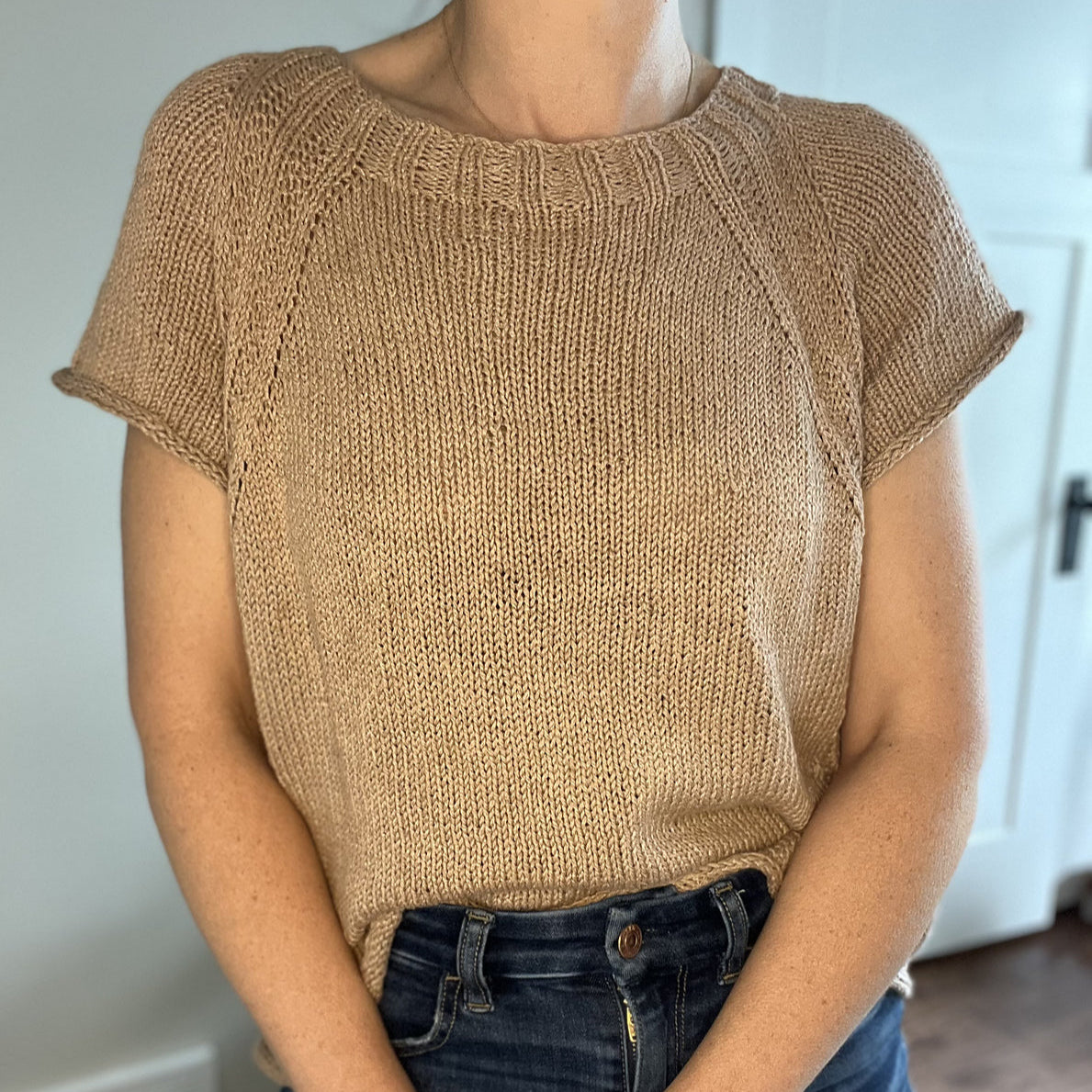 Woman wearing a beige short-sleeved knit sweater with ribbed neckline paired with dark jeans.