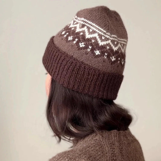 Person wearing a brown knitted hat with a pattern and matching sweater against a plain background