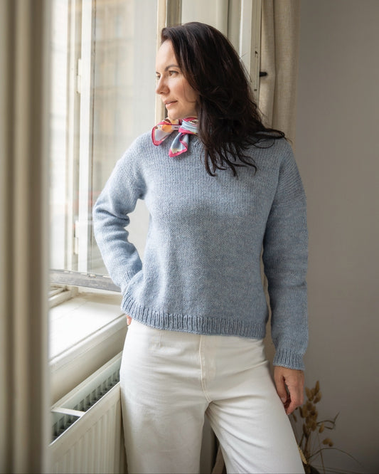 Woman wearing a blue sweater with a bow tie standing by a window.