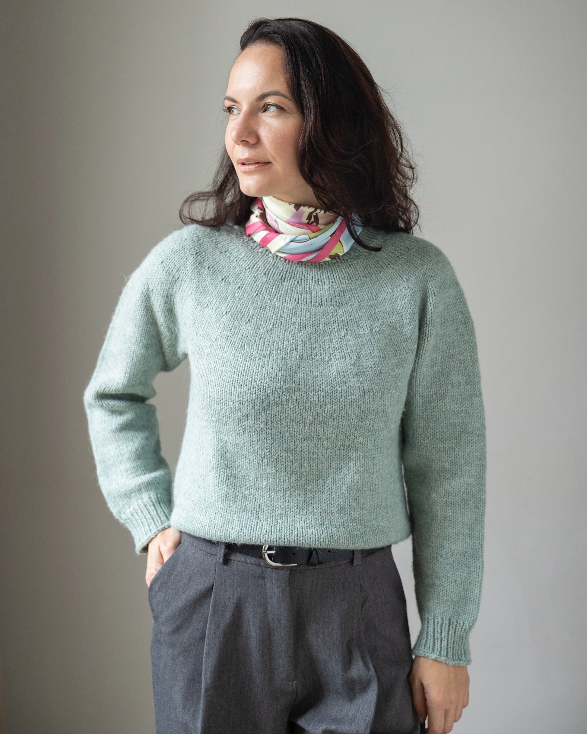 Woman wearing a light green sweater and dark pants against a plain background