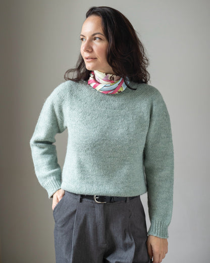 Woman wearing a light green sweater and dark pants against a plain background