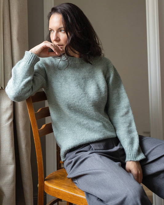 Woman wearing a light blue sweater and gray pants sitting on a wooden chair indoors.