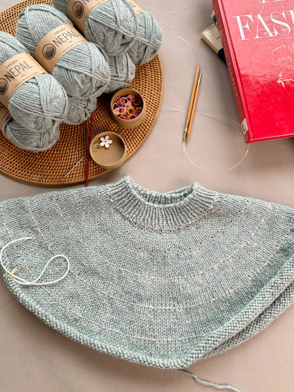 WIP First Star Sweater with knitted yoke on circular needles, surrounded by DROPS Nepal yarn, stitch markers, a flower-shaped progress keeper, knitting needles, and a red fashion book.