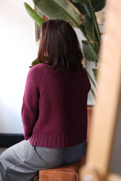 Back view of a woman wearing the Mellow Mood Cardigan in wine color, sitting on a brown sofa in a softly lit room with green plants in the background.