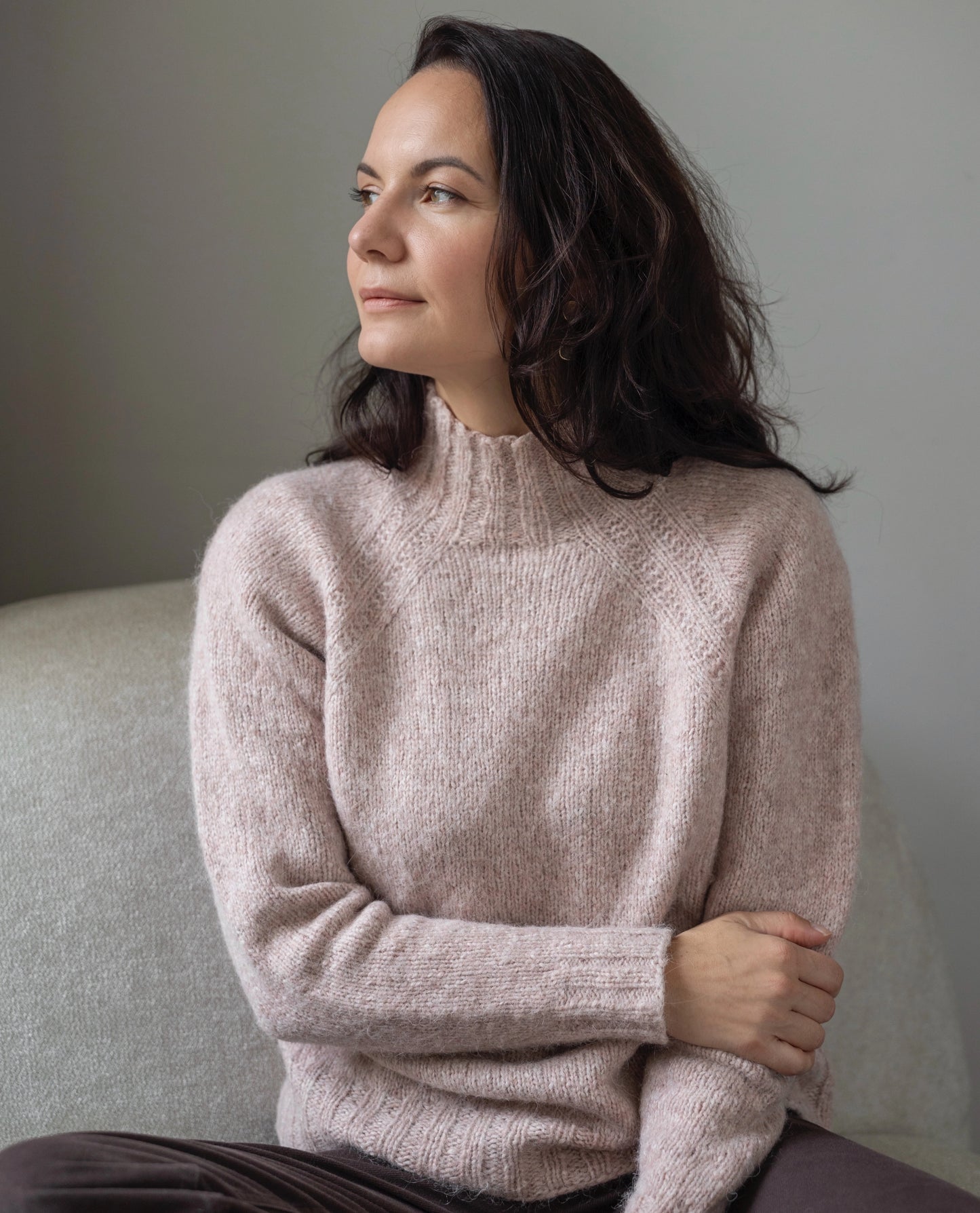 Woman wearing a light pink sweater sitting on a gray couch against a neutral wall.