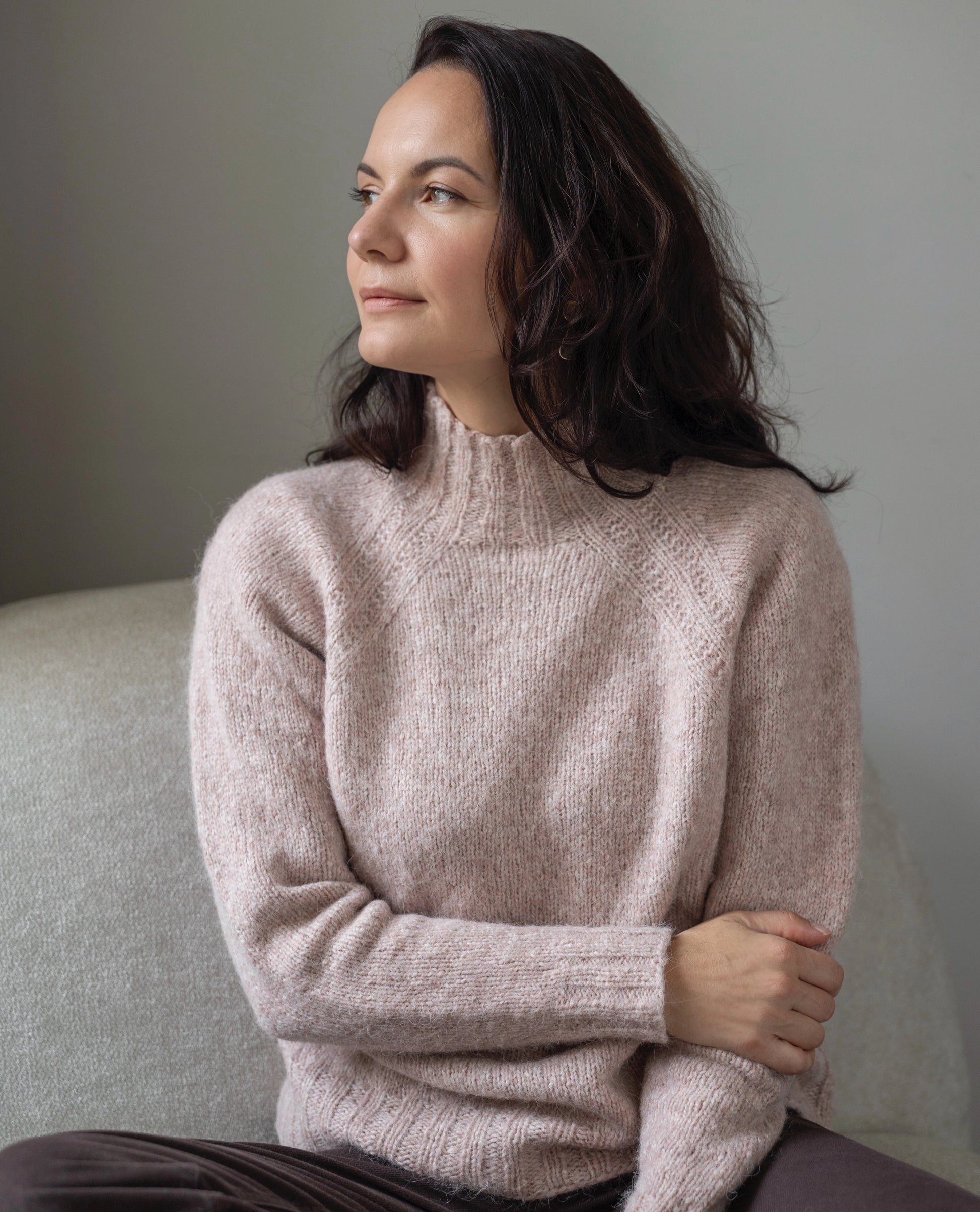 Woman wearing a light pink sweater sitting on a gray couch against a neutral wall.