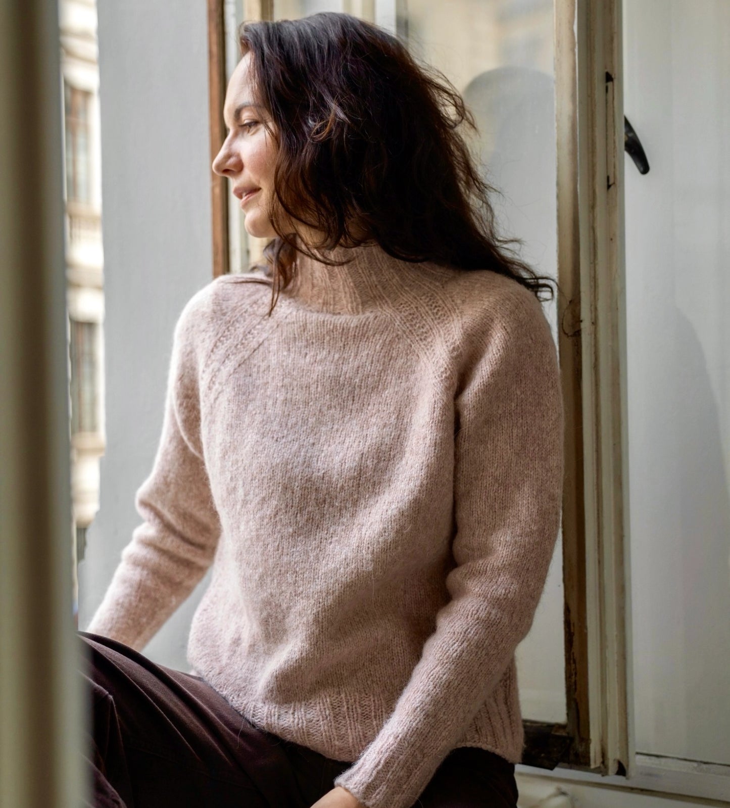 Woman wearing a light pink sweater sitting by a window.