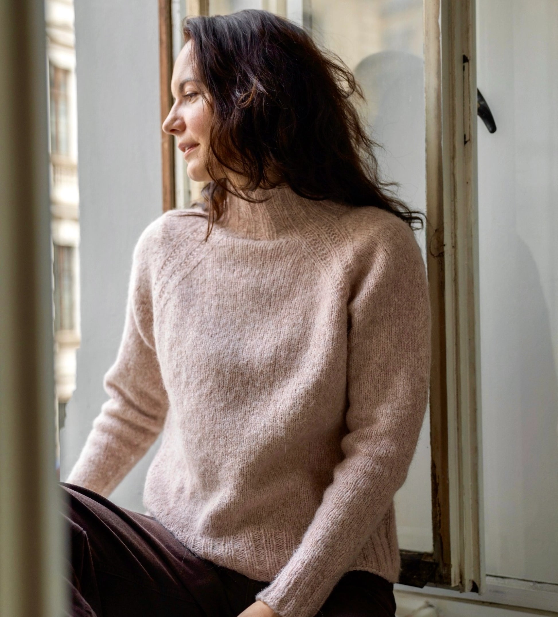 Woman wearing a light pink sweater sitting by a window.