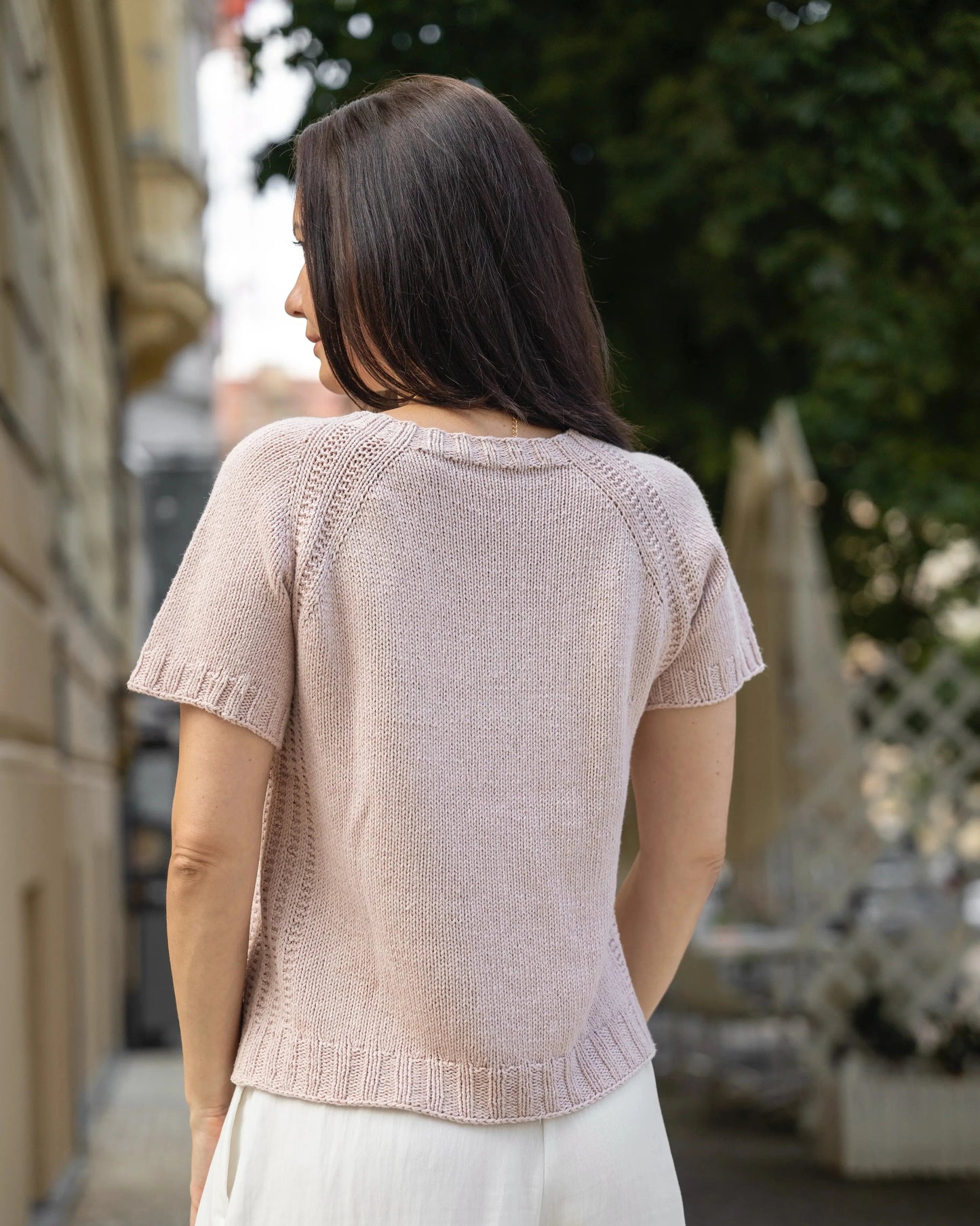 Woman wearing a light pink knitted top and white pants standing outdoors.