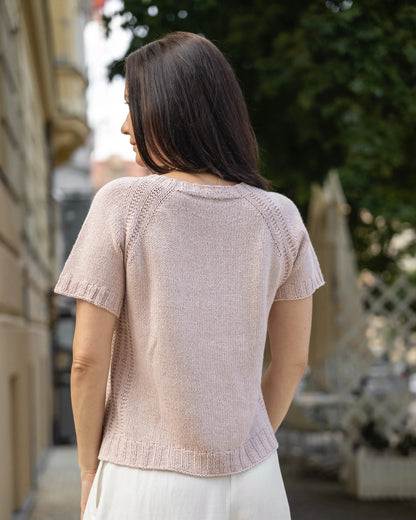 Woman wearing a light pink knitted top and white pants standing outdoors.