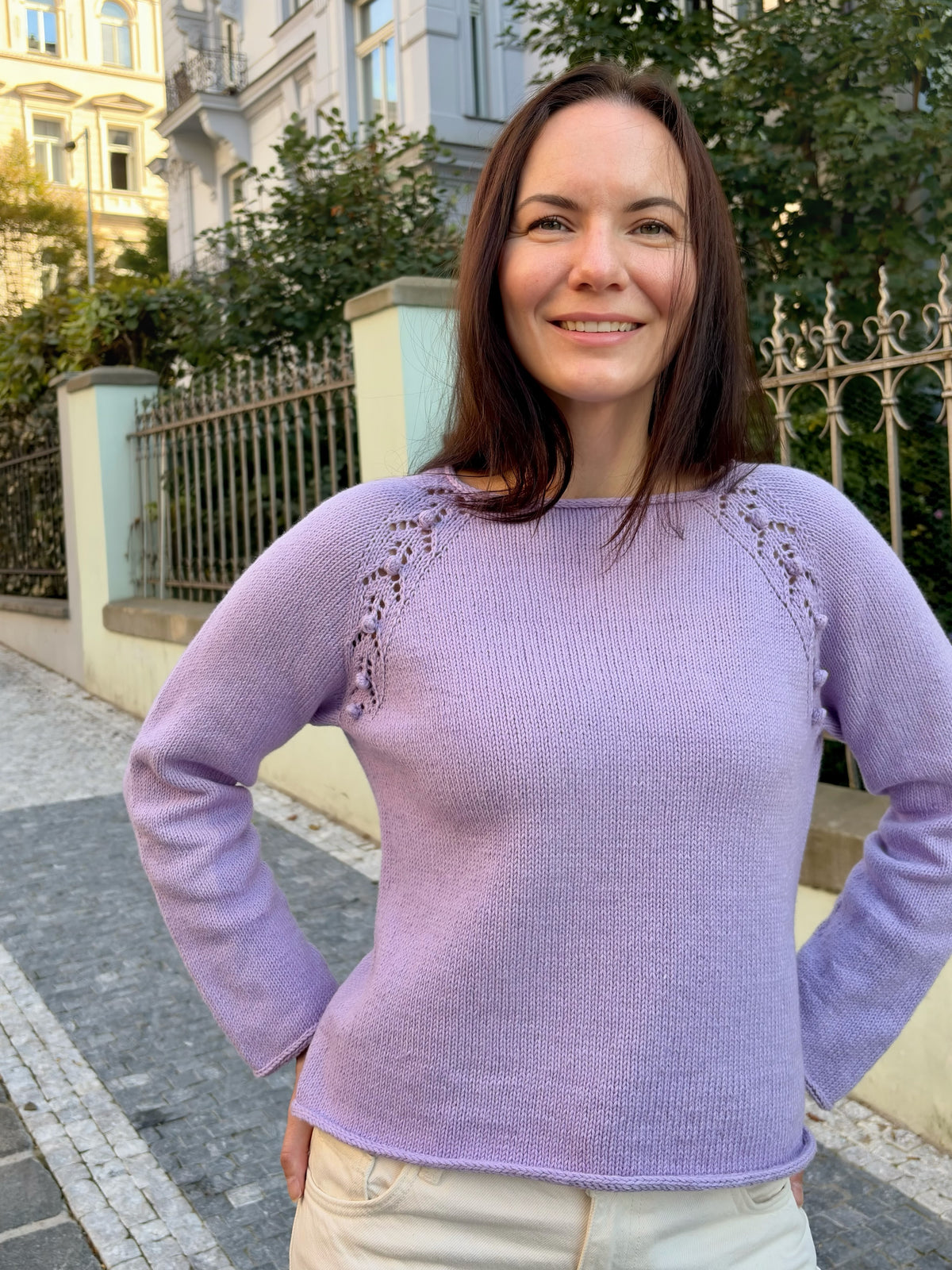 A woman wearing a light purple knit sweater with decorative lace-up details on the shoulders, standing outdoors on a paved street with buildings and a fence in the background.