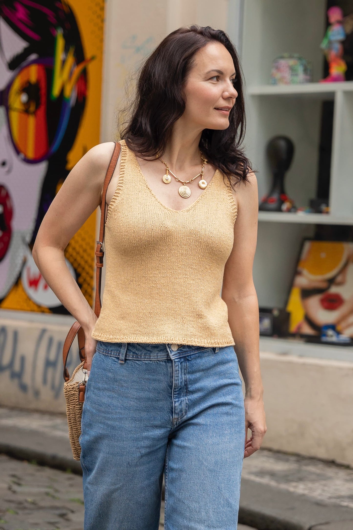 Woman wearing a yellow sleeveless top and blue jeans standing in front of colorful street art.