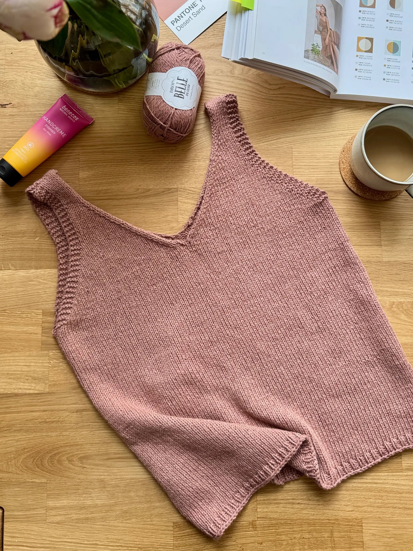 Knitted pink tank top on a wooden surface with yarn, coffee, and books in the background.