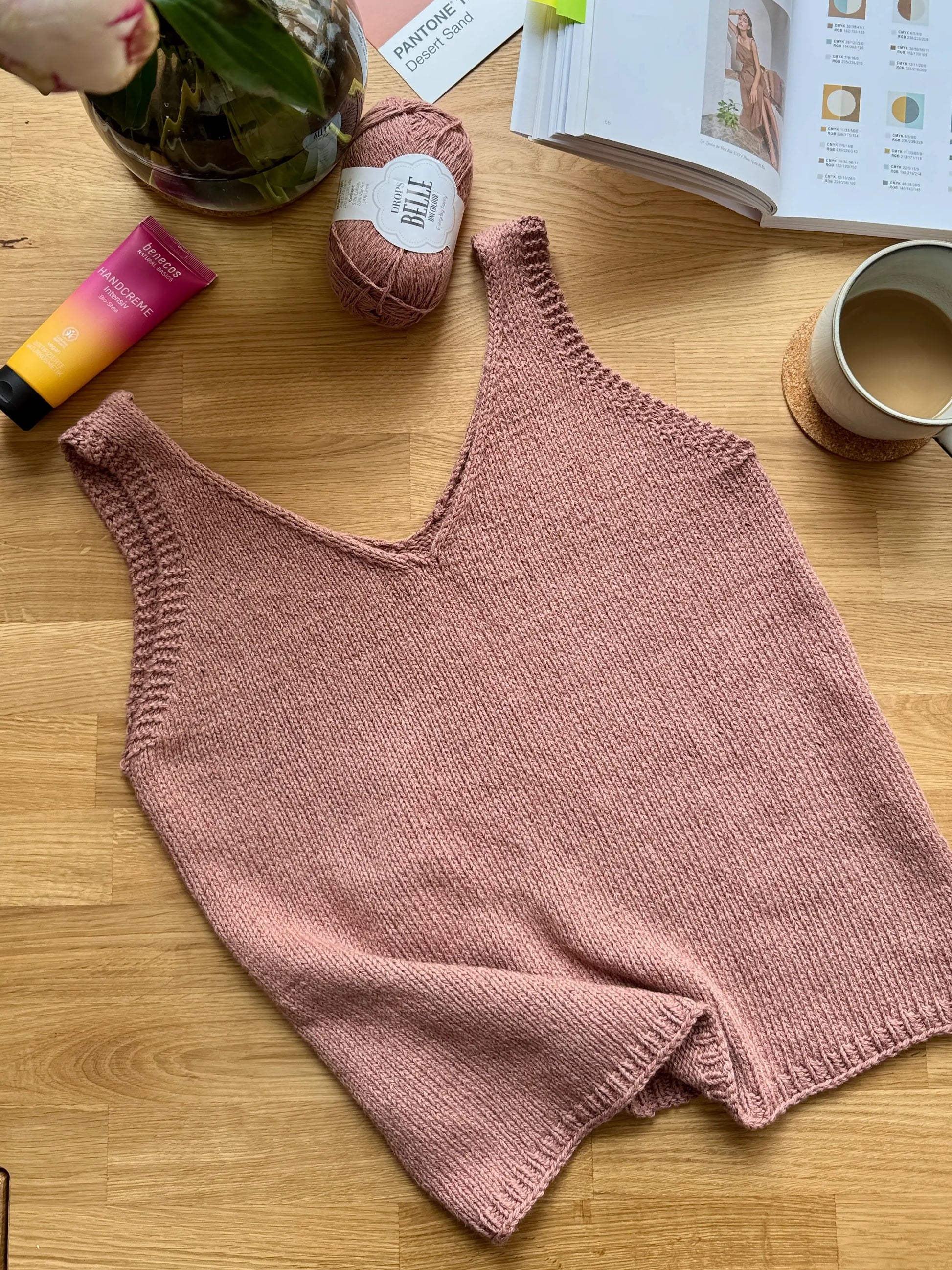 Knitted pink tank top on a wooden surface with yarn, coffee, and books in the background.