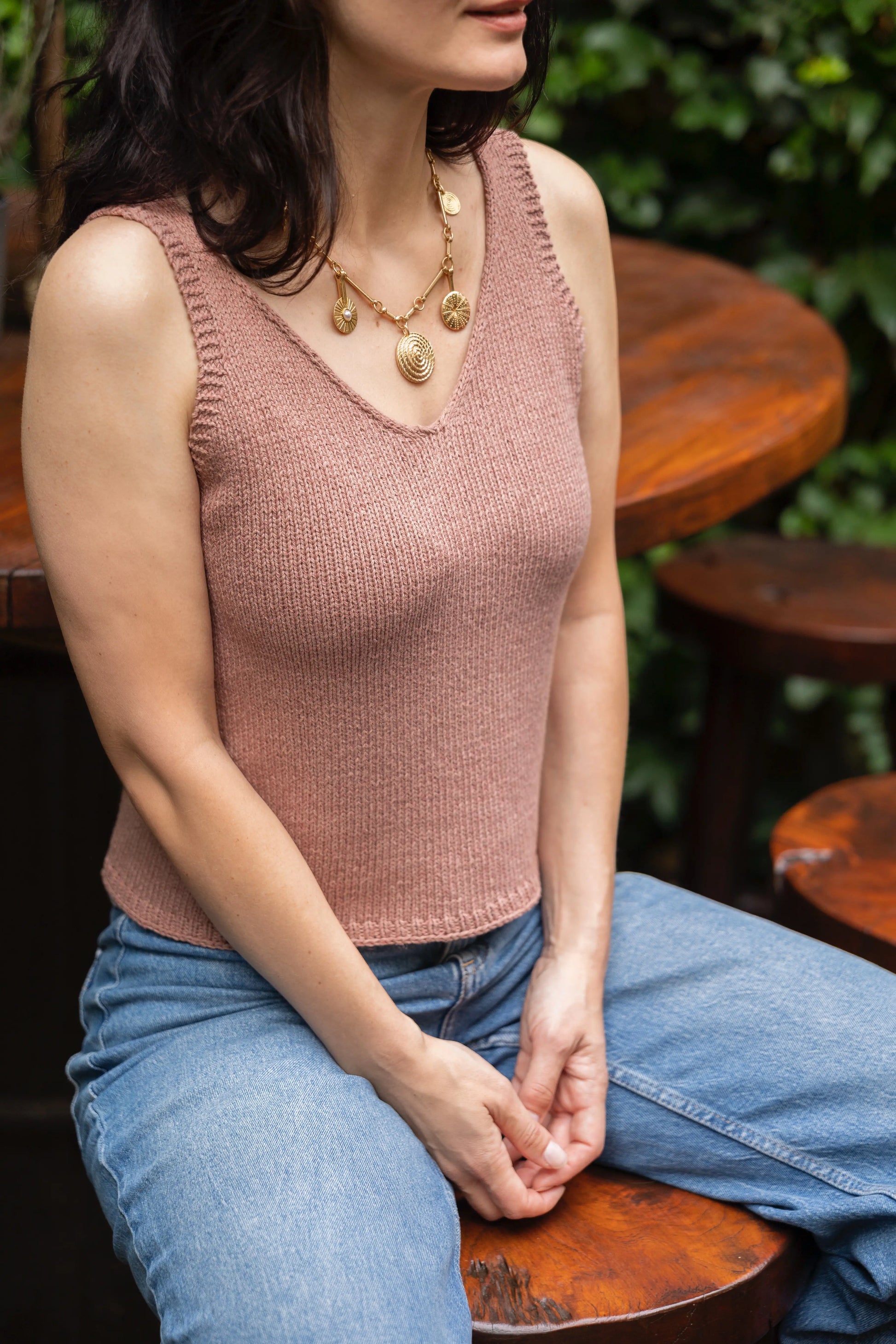 Woman wearing a pink knitted tank top and blue jeans sitting outdoors.