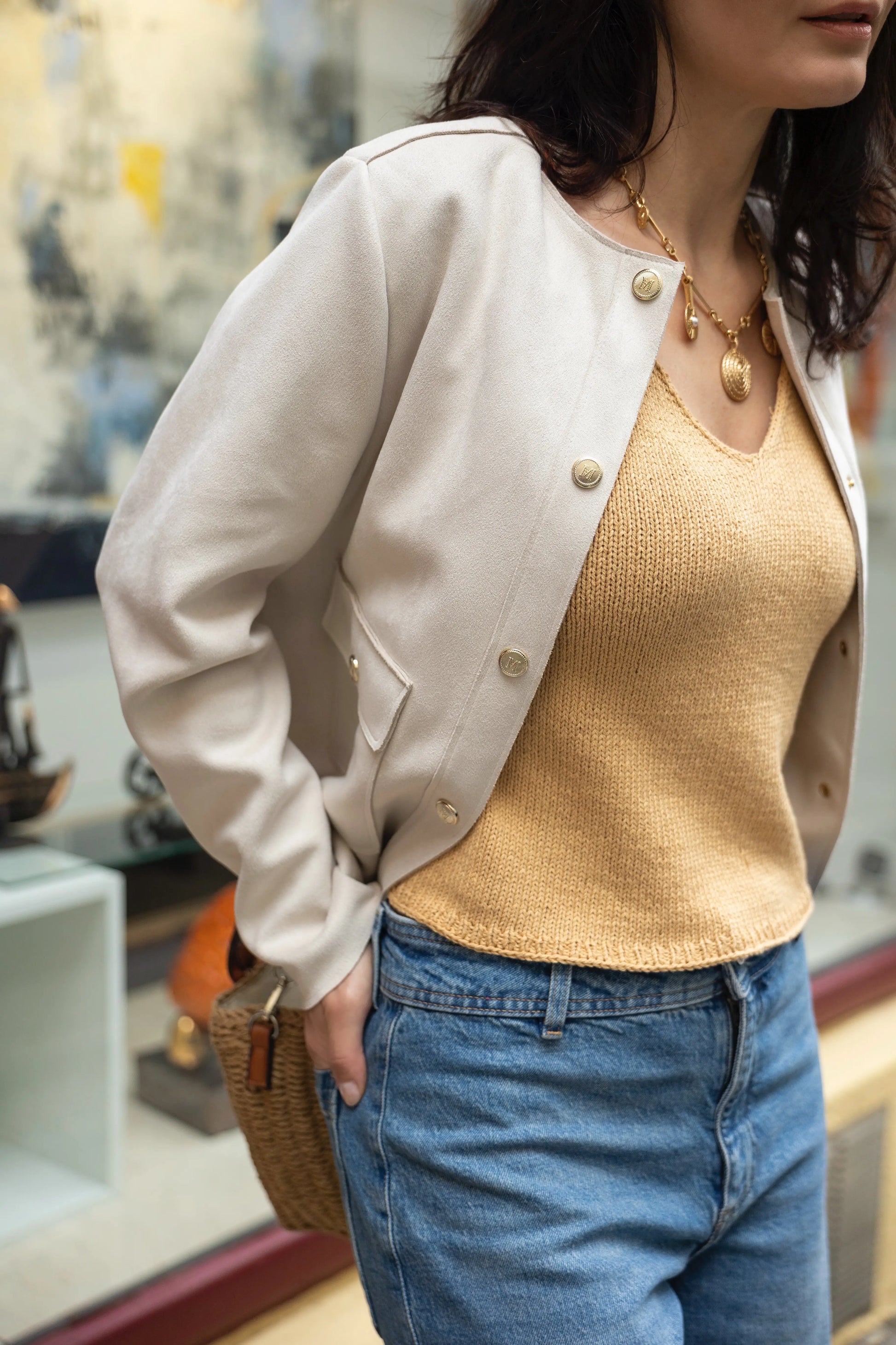 Woman wearing a beige jacket, gold top, and blue jeans with a blurred indoor background