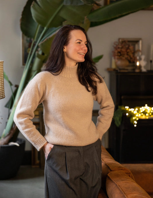 Woman in a beige sweater standing in a cozy living room with plants and a fireplace.