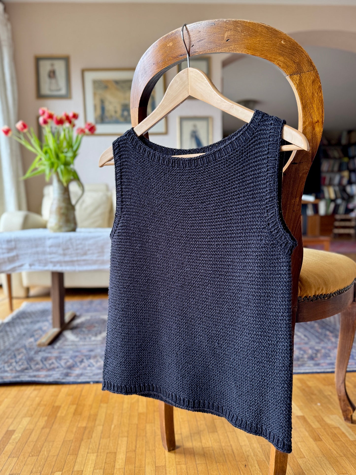 The Summer Soul Top in black, shown hanging on a wooden hanger to highlight its clean armholes, ribbed neckline, and seamless top-down construction. 