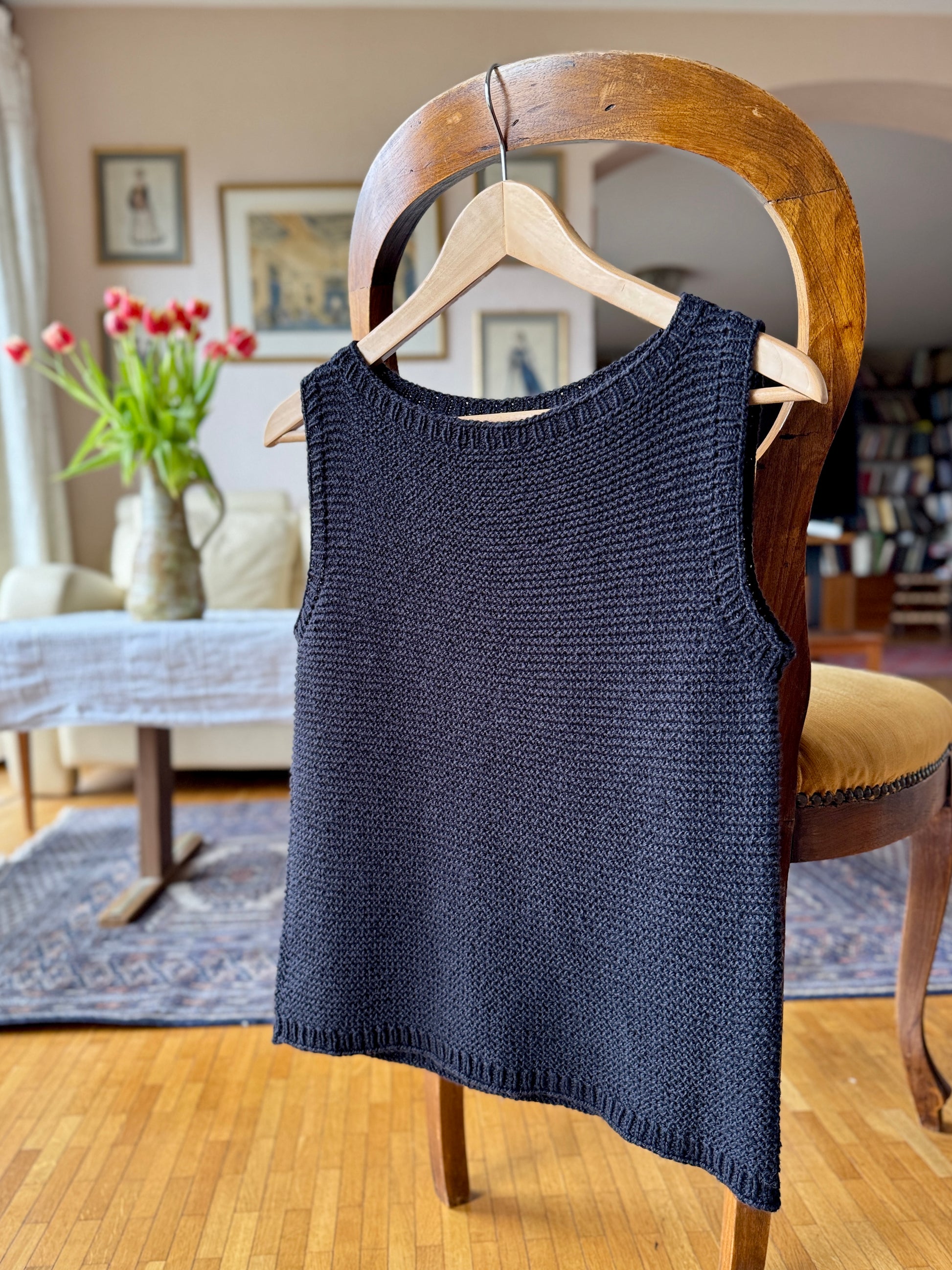 The Summer Soul Top in black, shown hanging on a wooden hanger to highlight its clean armholes, ribbed neckline, and seamless top-down construction. 